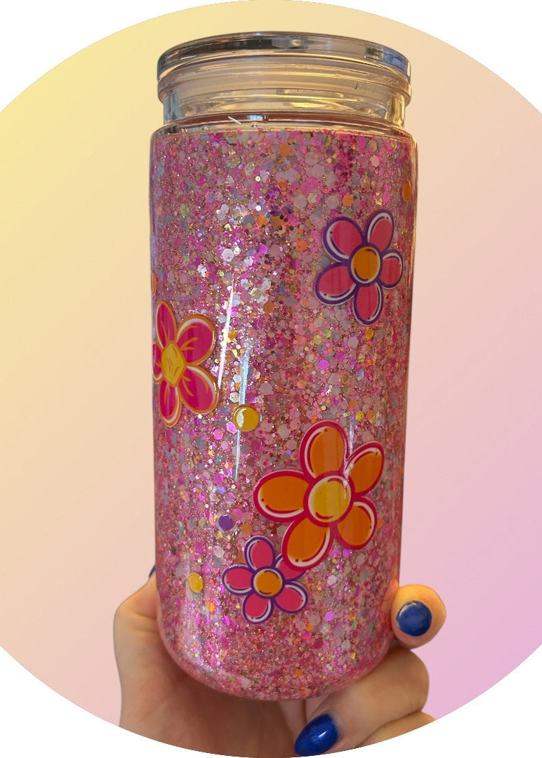 Pink glittery tumbler with floral designs held by a hand against a pink and yellow gradient background